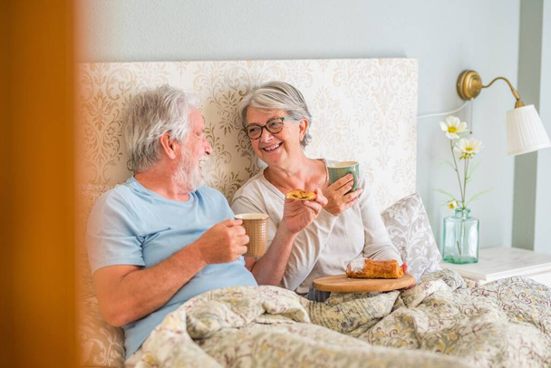 Старая старшая кавказская пара смеется и наслаждается завтраком в постели в спальне дома. Пожилая пара ест круассан и пьет кофе из чашки на завтрак дома   - Фото, изображение