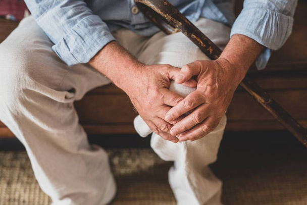 Close up of senior old man facing knee problem, sitting on sofa holding knee at home. Elderly man suffering from severe knee ache sitting with walking stick in living room.  - 写真・画像