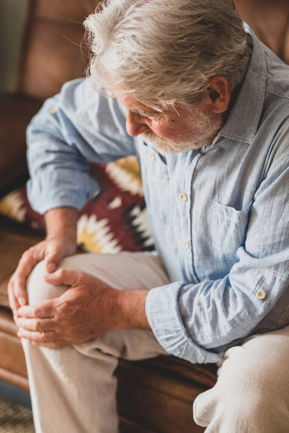Ανώτερος γέρος αντιμετωπίζει πρόβλημα γονάτου, κάθεται στον καναπέ κρατώντας το γόνατο στο σπίτι. Ηλικιωμένος άνθρωπος που υποφέρει από σοβαρό πόνο στο γόνατο κάθεται στο σαλόνι. Ηλικιωμένος άντρας αρπάζει το γόνατό του από πόνο   - Φωτογραφία, εικόνα