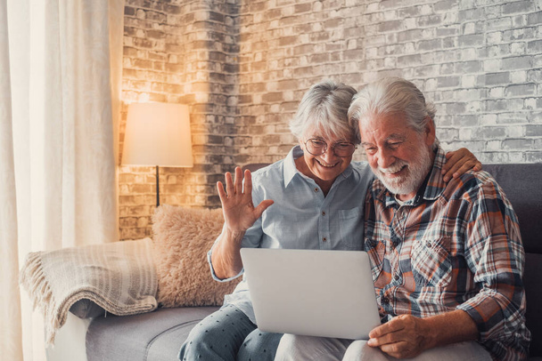 Симпатичная парочка пожилых людей, сидящих на диване, используя ноутбук вместе покупки и серфинга в сети. Два зрелых человека в гостиной наслаждаются технологиями, разговаривая на видео с друзьями или семьей.  - Фото, изображение