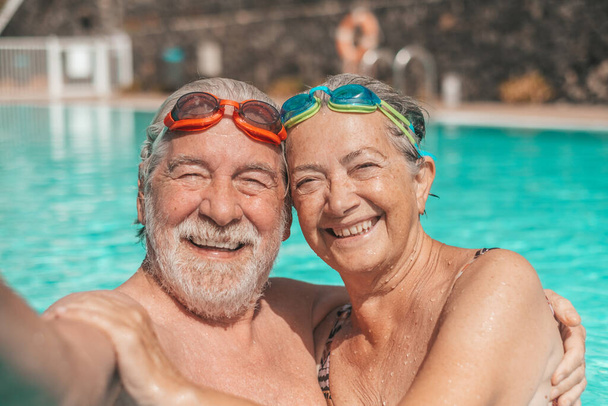 Pareja de dos felices seniors divirtiéndose y disfrutando juntos en la piscina tomando una foto selfie sonriendo y mirando a la cámara. Gente feliz disfrutando del verano al aire libre en el agua    - Foto, imagen