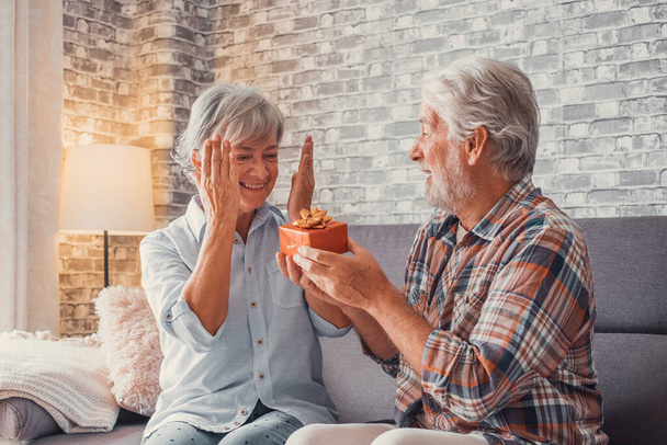 Portrait of two cute and old seniors at home having fun together. Mature man  giving a gift at his wife for Christmas or anniversary. Surprised pensioner woman looking at the present.  - Foto, imagen