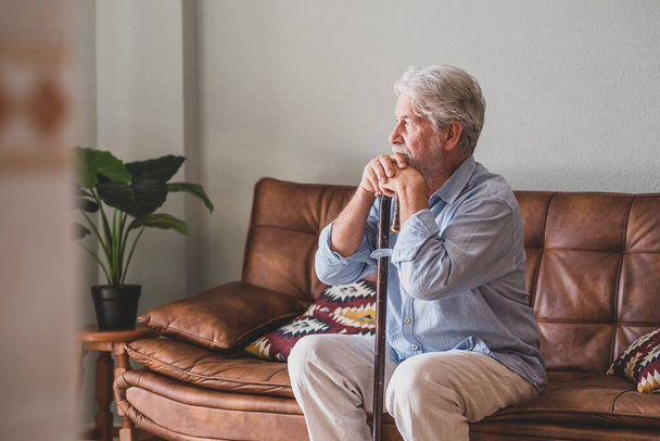 Задумчивый старший мужчина, сидя на диване, опирается на трость. Одинокий пожилой мужчина сидит дома в гостиной. Старик с седыми волосами, опирающийся на трость, сидит дома на диване  - Фото, изображение