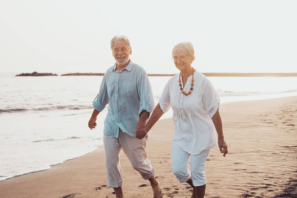 Ein Paar älterer, reifer Menschen, die zusammen auf dem Sand spazieren gehen und sich am Sand des Strandes amüsieren und den Moment genießen und leben. Zwei süße verliebte Senioren, die Spaß haben. Barfuß auf dem Wasser gehen - Foto, Bild