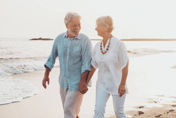 Couple of old mature people walking on the sand together and having fun on the sand of the beach enjoying and living the moment. Two cute seniors in love having fun. Barefoot walking on the water - Foto, imagen