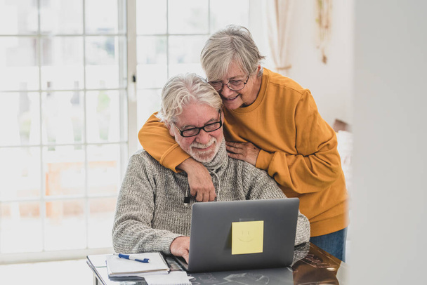 pari onnellista eläkeläistä työskentelee ja käyttää kannettavaa tietokonetta yhdessä kotona hymyillen ja pitäen hauskaa yhdessä - lukituselämäntapa  - Valokuva, kuva