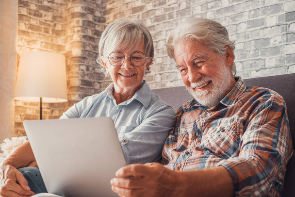 Cute couple of old people sitting on the sofa using laptop together shopping and surfing the net. Two mature people in the living room enjoying technology.  - Photo, Image