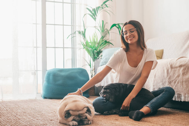 Portrait of beautiful woman in eyeglasses having fun with her pets pugs dogs sitting on floor in the living room of her house. Cheerful woman spending leisure time with her two cute pets dog at home  - Foto, afbeelding