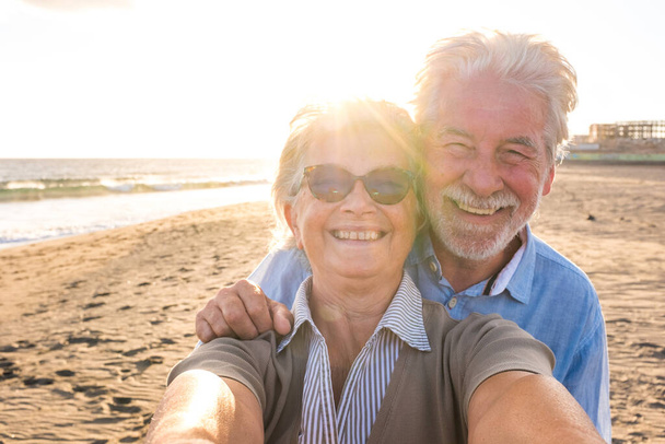 Porträt eines Paares reifer und alter Menschen, die den Sommer am Strand genießen und in die Kamera schauen, um ein Selfie mit dem Sonnenuntergang im Hintergrund zu machen. Zwei aktive Senioren im Freien unterwegs.  - Foto, Bild