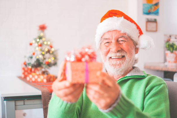 Rentner mit Nikolausmütze und warmer Kleidung feiert Weihnachten zu Hause. Der alte männliche Weihnachtsmann lächelt, während er ein Weihnachtsgeschenk in der Hand hält. Älterer Mann hält Weihnachtsgeschenkbox in der Hand   - Foto, Bild