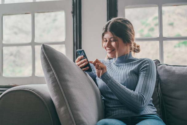 Nuori kaunis nainen istuu sohvalla kotona juttelemassa ja surffaamassa netissä. Naishenkilö hauskaa älypuhelimen verkossa. Muotokuva tyttö hymyilee käyttäen matkapuhelin tarkistaa sosiaalisen median  - Valokuva, kuva