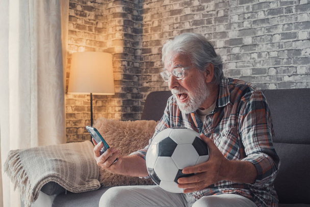 Portret van een gelukkige oude man die de overwinning viert van zijn liefdesteam voetbal of voetbal op de bank. Senior kijken wedstrijd op zijn telefoon winnen van een voetbalweddenschap.   - Foto, afbeelding