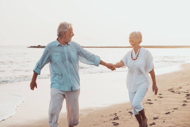 Pari vanhaa kypsää ihmistä kävelee hiekalla yhdessä ja pitää hauskaa rannalla nauttien ja eläen hetkestä. Kaksi söpöä senioria rakastuneena pitämässä hauskaa. Paljain jaloin kävellen veden päällä  - Valokuva, kuva