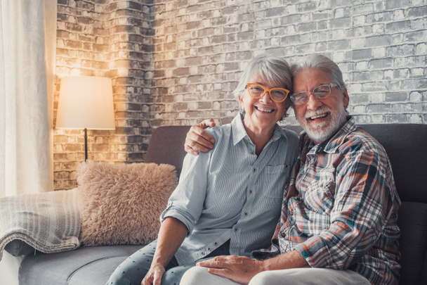 Carino e bella coppia di anziani sorridenti e guardando la fotocamera divertirsi a casa insieme. Ritratto di anziani che indossano occhiali seduti sul divano godendo e rilassandosi.  - Foto, immagini