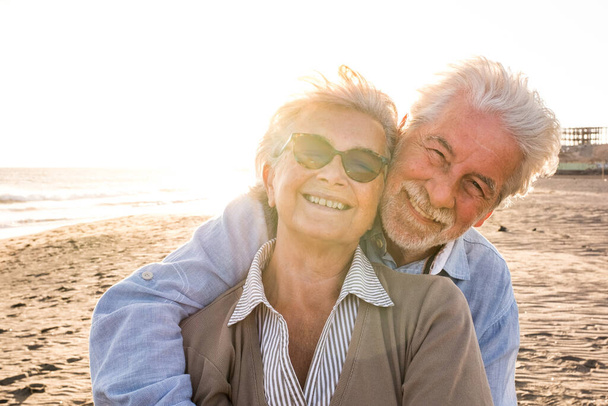 Portret pary dojrzałych i starszych ludzi cieszących się latem na plaży patrzących w stronę aparatu uśmiechniętego i bawiących się razem z zachodem słońca w tle. Dwóch aktywnych seniorów podróżujących na zewnątrz.  - Zdjęcie, obraz