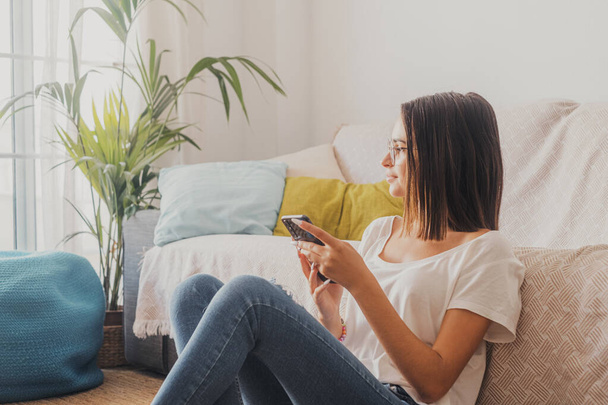 Poważna kobieta w okularach SMS-uje przez telefon komórkowy siedząc na podłodze w salonie swojego domu. Piękna pani spędza wolny czas korzystając ze smartfona w domu  - Zdjęcie, obraz