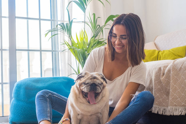 Portrait of beautiful woman in eyeglasses having fun with her pet pug dog sitting on floor in the living room of her house. Cheerful woman spending leisure time with her cute pet dog at home   - Photo, Image