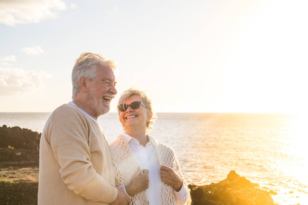 Nahaufnahme und Porträt von zwei glücklichen und aktiven Senioren oder Rentnern, die Spaß haben und den Sonnenuntergang lächelnd mit dem Meer betrachten - alte Menschen im Freien, die gemeinsam Urlaub machen  - Foto, Bild