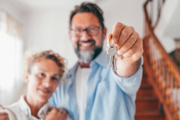 Porträt eines glücklichen kaukasischen Paares mit neuen Hausschlüsseln. Freudige Frau und Mann, die sich über den Kauf eines neuen Eigenheims freuen. Stolze Hausbesitzer stehen nach Umzug drinnen und zeigen Schlüssel.  - Foto, Bild