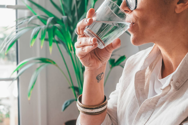Close up of young woman drinking fresh glass of water in domestic room at home. Close up cropped of caucasian thirsty female holding transparent glass drinks purified water at house - Photo, Image