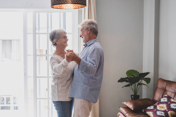 Romantický milující starší pár držící se za ruce a užívající si tance v obývacím pokoji domu, starší šťastný pár oslavující tancem doma. Starý manžel a manželka baví čas v moderním bytě  - Fotografie, Obrázek