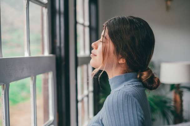 Одна молодая женщина смотрит в окно, веселясь и расслабляясь пасмурный день дома одна. Женщина чувствует себя хорошо в помещении.  - Фото, изображение