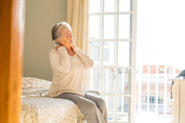Idős nő szenved a nyak fájdalom alvás után, dörzsölés izmok, idős nő ül az ágyon megható nyak érzés kellemetlen, mert kényelmetlen ágy otthon  - Fotó, kép