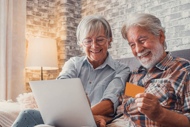 Portret pary uroczych dwóch szczęśliwych i podekscytowanych seniorów kupujących i wydających pieniądze na zakupy online z laptopem na sofie w domu. Korzystanie z karty kredytowej razem zakup w Internecie.  - Zdjęcie, obraz