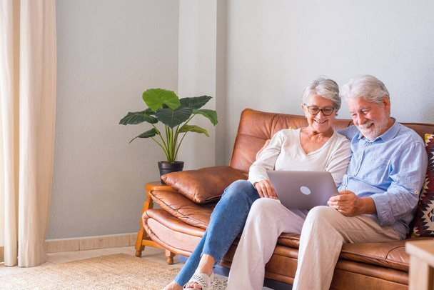 Fröhliches Seniorenpaar mit Laptop auf dem Sofa sitzend und lächelnd. Älteres glückliches Paar entspannt beim Surfen auf dem Laptop im Wohnzimmer sitzend. Altes Ehepaar schaut Medieninhalte mit Laptop   - Foto, Bild