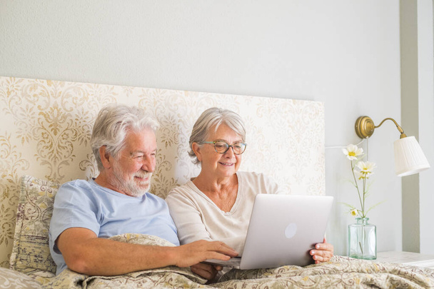 Улыбающаяся пожилая пара просматривает и смотрит социальные сети с помощью ноутбука в постели дома. Расслабленный старый муж и жена проводят досуг в постели, делясь онлайн фильмом в спальне в современных апартаментах  - Фото, изображение