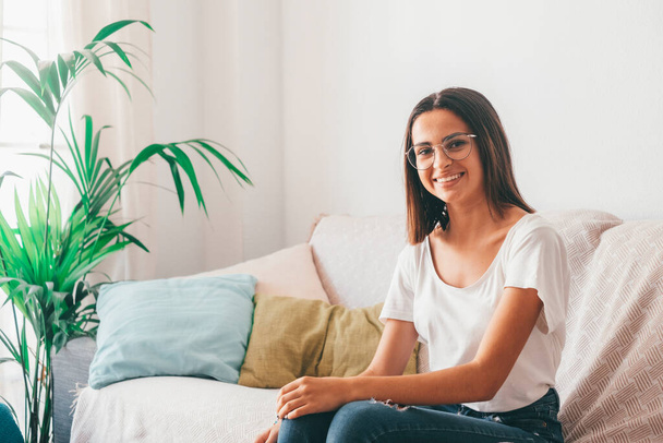 Happy young woman in eyeglasses relaxing on sofa in the living room of her house. Beautiful satisfied lady sitting on couch at modern apartment. Pretty young female looking at camera while sitting at home  - Photo, Image