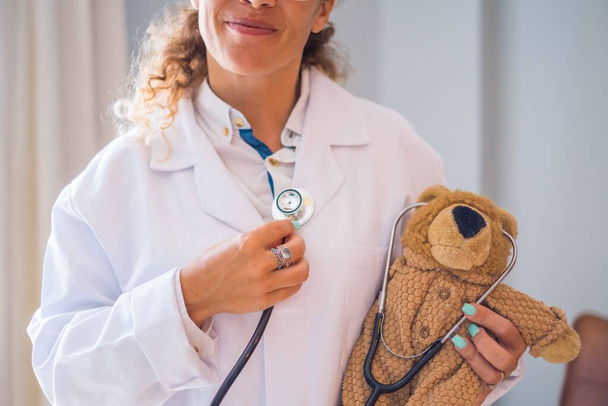 Молодая женщина-врач в форме и проверяет себя стетоскопом плюшевого мишки в клинике. Крупный план врача или медицинского работника, развлекающегося с Тедди в больнице    - Фото, изображение