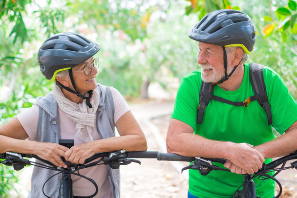 Portrait de couple de personnes âgées et heureuses amoureuses qui se regardent en souriant et en s'amusant avec leurs vélos dans la nature à l'extérieur ensemble se sentant bien et en bonne santé.  - Photo, image
