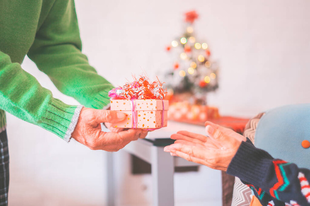 Hands of senior man giving surprise Christmas gift box to his old wife. Loving old heterosexual couple celebrating Christmas festival together at home. Couple exchanging gift in front of Christmas tree   - Foto, afbeelding