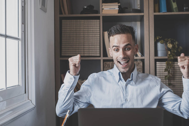 Şaşırmış Kafkasyalı genç işadamı ofisteki dizüstü bilgisayarla online olarak iyi haberler alarak başarıyı kutluyor. İş yerindeki başarıdan memnun olan erkek işletme yöneticisi - Fotoğraf, Görsel