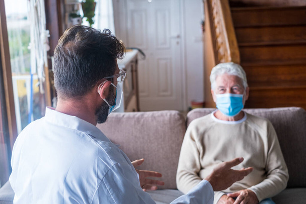 Male professional doctor consulting senior patient during medical care visit wearing masks. Young man physician and old mature senior talking providing medical assistance sitting on sofa. Elderly people home care. Covid and coronavirus positive test - Photo, Image