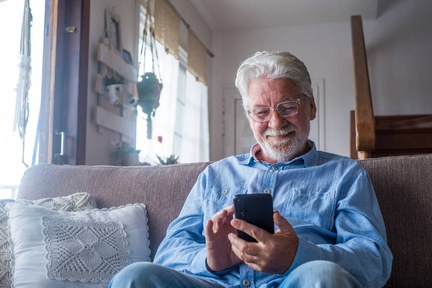 Γέρος χαμογελώντας κάθεται στον καναπέ στο σαλόνι κρατώντας τηλέφωνο, απολαμβάνοντας τη χρήση smartphone αίσθημα ικανοποιημένοι στέλνοντας μηνύματα, καλώντας τους φίλους, σερφάροντας web online έννοια   - Φωτογραφία, εικόνα