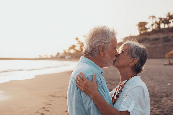 Κοντινό πλάνο από δύο χαριτωμένους και χαρούμενους ηλικιωμένους που διασκεδάζουν και απολαμβάνουν μαζί μια μέρα ηλιοβασιλέματος στην παραλία. Ώριμο ζευγάρι ερωτευμένο φιλιέται μαζί με το ηλιοβασίλεμα στο παρασκήνιο. - Φωτογραφία, εικόνα