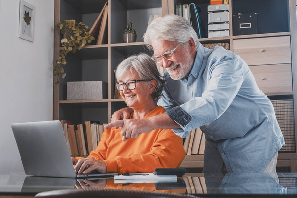 Elderly man and woman having fun using laptop at home office. Smiling old husband pointing finger and showing something online to wife typing on keyboard. Senior couple watching media content and enjoying at home  - Foto, Bild