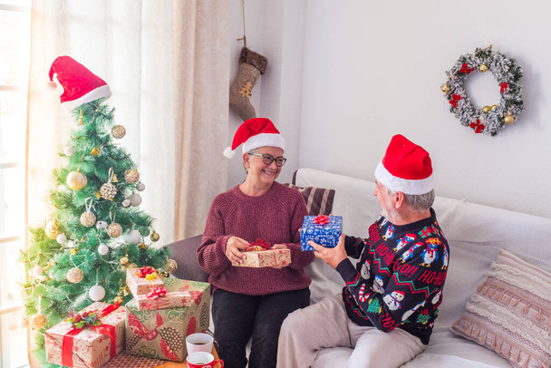 Senior couple in warm clothing and Santa hat holding each others hands in front of decorated Christmas tree at home. Loving old romantic heterosexual couple celebrating Christmas festival together  - Photo, Image