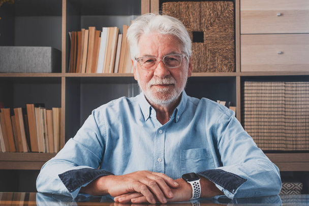 Portrait of smiling senior man in eyeglasses and gray hair looking at camera in front of book shelf. Happy old online male tutor giving educational training class lecture or doing video conference call - Photo, Image