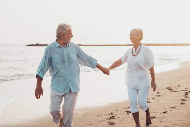 Couple of old mature people walking on the sand together and having fun on the sand of the beach enjoying and living the moment. Two cute seniors in love having fun. Barefoot walking on the water  - Photo, Image
