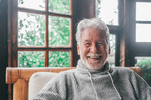 Портрет старика, улыбающегося и смотрящего на камеру, сидящую на диване из дома. Крупный план мужчина пожилой веселый в помещении.  - Фото, изображение