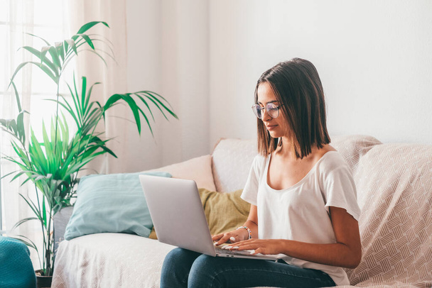 Szczęśliwa młoda kobieta w okularach, pisząca na laptopie siedząc na kanapie w salonie swojego domu. Piękna pani spędza wolny czas korzystając z laptopa. Wolny strzelec pracujący w domu  - Zdjęcie, obraz
