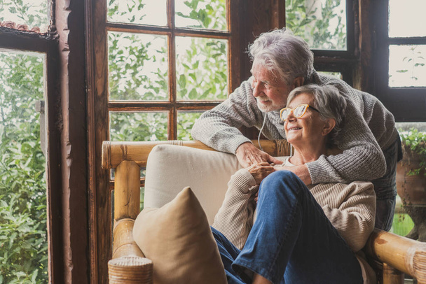 Old caucasian couple spending leisure time looking out through window at home. Loving husband embracing wife from behind sitting on armchair at house and admiring view from transparent door glass.  - Photo, Image
