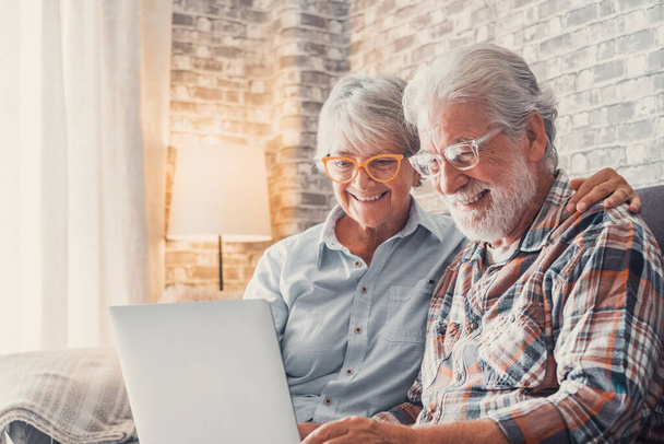Roztomilý pár starých lidí sedí na pohovce pomocí notebooku spolu nakupování a surfování na internetu. Dva dospělí lidé nosí brýle v obýváku a užívají si technologie. Portrét zamilovaných seniorů.   - Fotografie, Obrázek