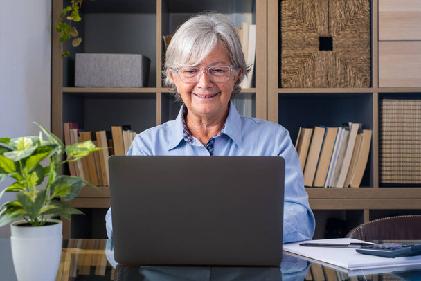 Щаслива стара кавказька бізнес-леді посміхається робочий онлайн-перегляд веб-сеансу подкасту на ноутбуці та навчальному курсі конференції з викликом ноутбуків робить нотатки сидять за робочим столом, концепція електронного навчання
  - Фото, зображення