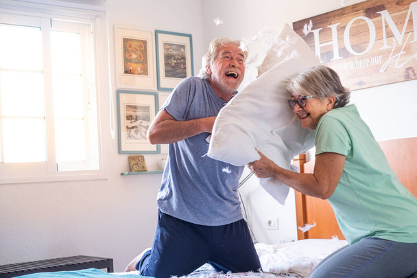 2人の幸せな高齢者が枕を楽しんで家の戦いでベッドで遊ぶのを楽しんでいます-朝の枕戦争屋内  - 写真・画像