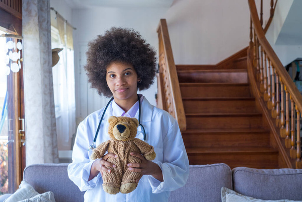 Portret młodej afrykańskiej pielęgniarki w mundurze medycznym trzymającej pluszową zabawkę pokazującą miłość do małej pacjentki, szczęśliwej kobiety lekarz pediatra pozujący z misiaczkiem w szpitalu lub klinice  - Zdjęcie, obraz