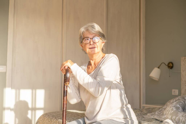 Thoughtful senior woman leaning on walking stick while sitting on bed. Lonely elderly woman sitting in bedroom at home. Old woman enjoying morning sunlight at home  - Foto, Bild